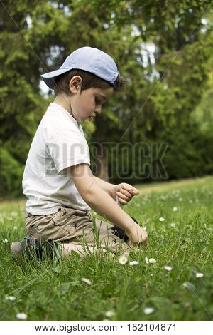 Playing boy on lawn. Funny white child with baseball cap. Young lad sits in grass. Youngster with thoughtful face. In captivity of activity. Son spends leisure time activities in house garden.