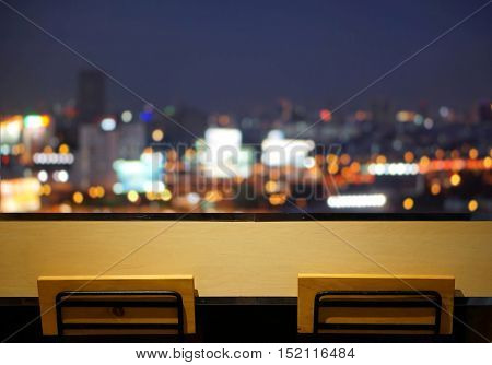 wooden long bar table top with 2 simple chairs at window  with blurry cityscape bokeh light in the dark as background