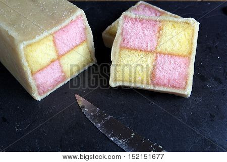 Freshly Cut Battenberg Cake On Dark Slate Background With Knife. Pink And Yellow Sponge Covered In M