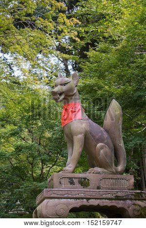 Kyoto Japan - September 17 2016: Fushimi Inari Taisha Shinto Shrine. Kitsune the brown fox messenger and server of God Inari holds a magic ball in its mouth and sports a red apron. Against green tree foliage.
