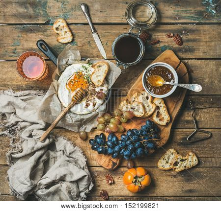 Cheese, fruit and wine set. Camembert in small pan, honey, fig jam, persimmon, grapes, pecan nuts, grilled baguette slices and glass of rose wine over rustic wooden background, top view