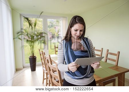 Beautiful young mother with her newborn baby son in sling at home, holding tablet, reading something