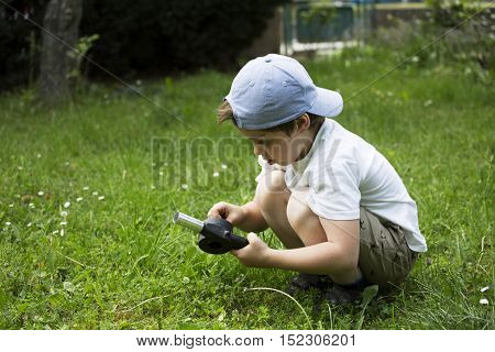 Playing boy on lawn. Funny white child with baseball cap. Young lad sits in grass. Youngster with thoughtful face. In captivity of activity. Son spends leisure time activities in house garden.