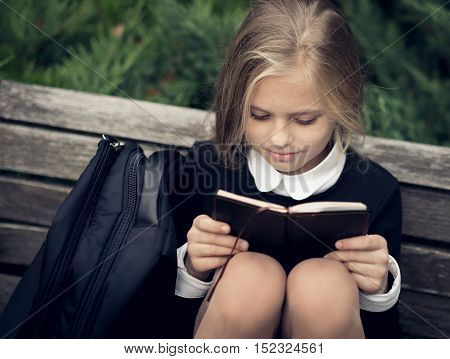 Beautiful blond girl in a school uniform sits on a park bench and reading a book. School uniform.Education