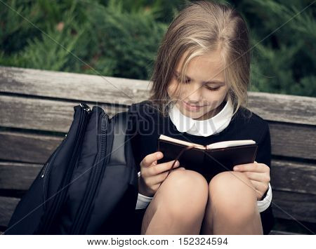 Beautiful blond girl in a school uniform sits on a park bench and reading a book. School uniform.Education