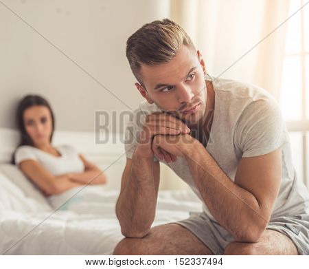 Young couple having a quarrel. Handsome young man is sitting in the foreground while his wife is lying in bed