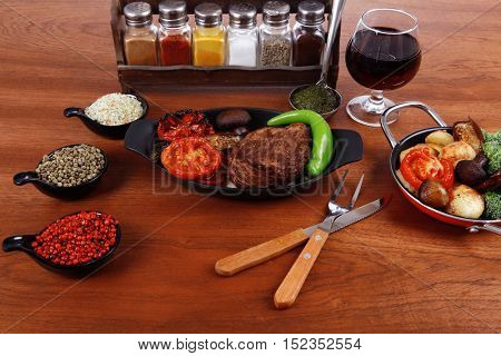 fresh roast beef meat rib eye steak with tomatoes,potatoes, hot chili pepper gern and pink peppercorn saucer, cutlery, set of spice, and red wine glass on light walnut table