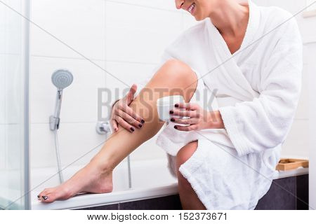 Woman in bathroom sitting at edge of bathtub putting on lotion on legs