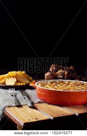 Wooden table served for Thanksgiving dinner. Close view at pumpkin pie. Chips, chicken legs, and mash potatoes on a black background. Vertical shot. Dark photo