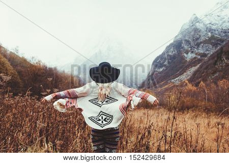Boho woman wearing hat and poncho standing by the mountain. Cold weather, snow on hills. Winter hiking. Wanderlust.