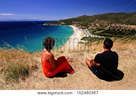 Man and woman in orange dress sitting and enjoying view on beautiful beach and blue sea with transparent water in Sarinia Italy. The beach is called Solanas Sinnai.