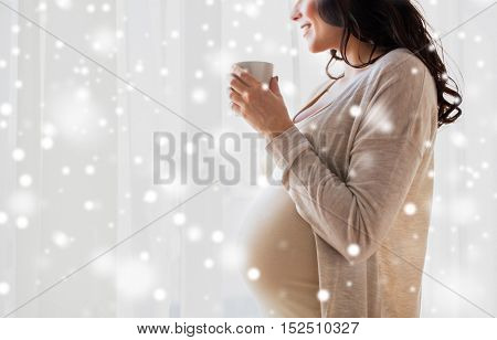 pregnancy, people, winter, christmas and expectation concept - close up of happy pregnant woman with cup drinking tea looking through window at home over snow