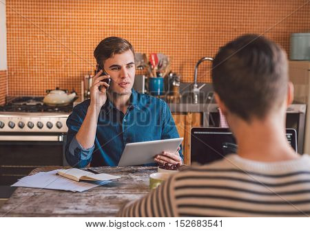 Young business owners sitting at a kitchen table at home using a cellphone, laptop and digital tablet while working together on their home based business