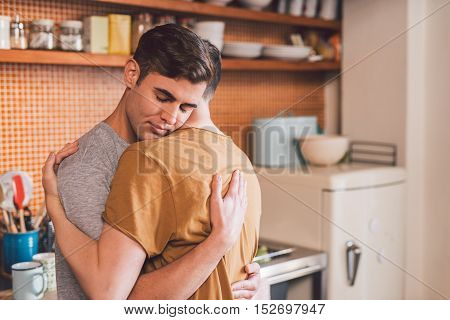 Affectionate young gay couple hugging each other with their eyes closed while standing in their kitchen in the morning