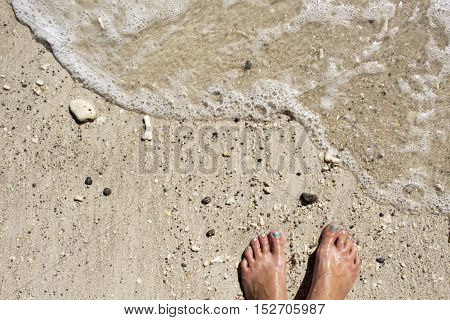 Woman's feet near wave on white sand beach. Sunny afternoon at the tropical island. Barefoot on seashore background image with place for text. Optimistic horizontal picture from summer vacation