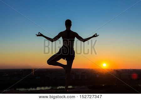 Silhouette of sporty woman practicing yoga in the park at sunset - half lotus tree pose. Sunset light, golden hour. Freedom, health and yoga concept