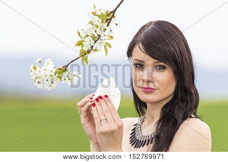 Sad hayfever girl in spring nature with blossom branch season. The beautiful young woman is pained by her allergy every year. She holds a tissue in her hands.