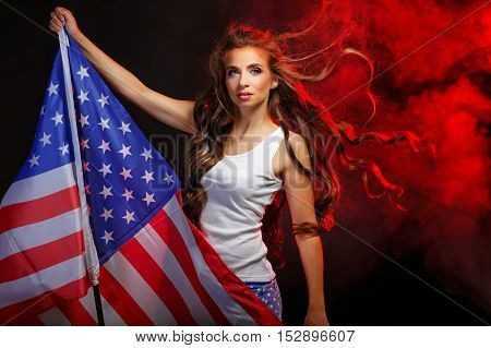 Young attractive girl holding a US flag on the flagpole. She a T-shirt and shorts with a print of the flag. Long curly hair flying. Portrait of a real patriot. Stars and Stripes. Smoke in the background.