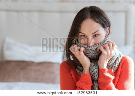 Keeping warmth. Good looking nice brunette woman standing in the room and looking at you while wearing a warm knitted scarf