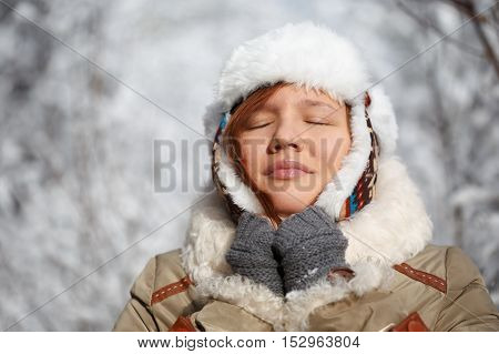 Beautiful young smiling woman in gray fingerless knitted mittens cap with ear flaps and coat with white fur at the background of snowy forest.