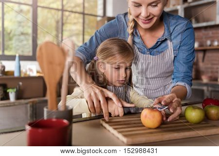 Little mothers helper. Joyful cheerful involved girl cutting apples with her mother while sitting at the table and cooking in the kitchen