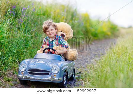 Little preschool kid boy driving big toy car and having fun with playing with his plush toy bear, outdoors. Child enjoying warm summer day in nature landscape