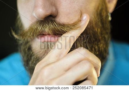 Bearded Man In A Blue Denim Shirt Twirling His Mustache With His Fingers. Portrait Of A Hipster On B