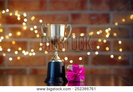 Handmade Gift Box With Purple Bow And Golden Prize Cup On Wooden Table With Fairy Lights On Bokeh Ba