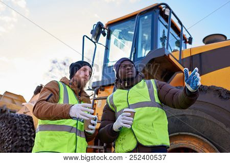 Low Angle Portrait Of Two Workers, One African-american, Drinking Coffee And Chatting Standing Next 