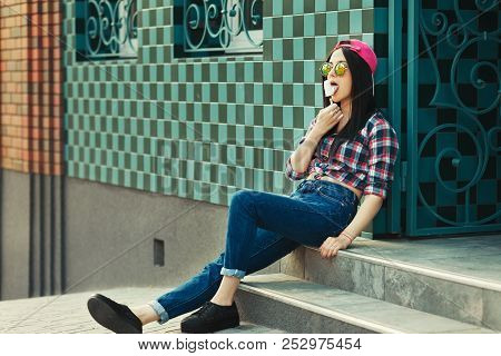 Young Hipster Style Girl Outdoor In Creative Sunglasses And Eating Ice Cream On Green Urban Backgrou