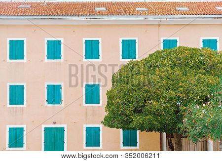 The House Has A Red Tiled Roof And Closed Turquoise Shutters In The Windows. Flowering Tree In The F