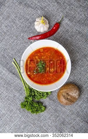 Beef Goulash Soup With Tongue. A White Oval Plate With Red Soup Stands On A Gray Canvas. Next To It 