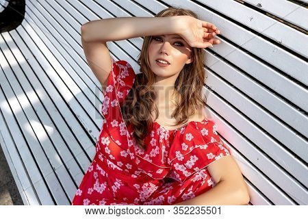 Beautiful Young Girl, Model, Brown-haired Woman With Long Hair In A Red Dress Sits On A Bench And Po