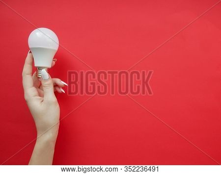 Female Hand Holds An Led Lamp On A Red Background. Energy Saving Concept, Alternative Energy Sources