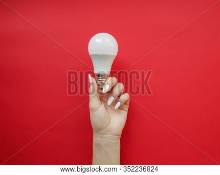 Female Hand Holds An Led Lamp On A Red Background. Energy Saving Concept, Alternative Energy Sources