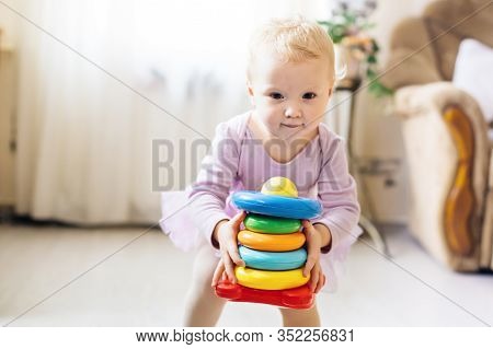 Girl Plays Logical Pyramid On Floor In Living Room On Sunny Day. Montessori Wooden Toy Folded Pyrami