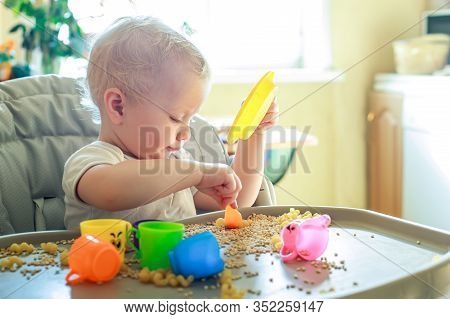 Small Kid Sits On Feeding Chair And Plays With Toy Utensils And Grain. Development Of Fine Motor Ski