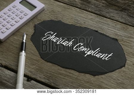 Calculator, Pen And A Piece Of Black Paper Written Shariah Compliant On Wooden Background.