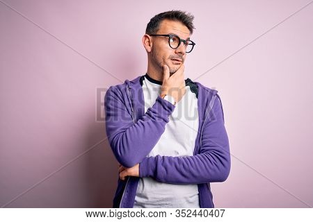 Young handsome man wearing purple sweatshirt and glasses standing over pink background Thinking worried about a question, concerned and nervous with hand on chin