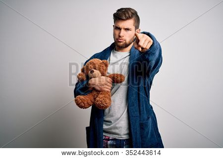 Young blond man with beard and blue eyes wearing pajama holding teddy bear pointing with finger to the camera and to you, hand sign, positive and confident gesture from the front