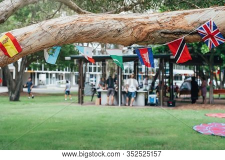 Flags Of Different Countries In The Tree Branch With Blured People In Park Doing Barbecue Together. 