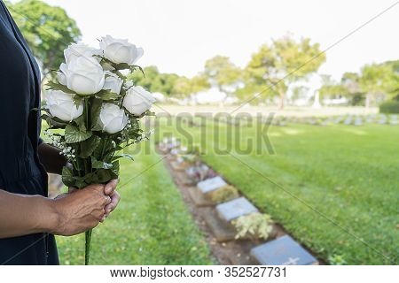 Mourning Young Woman Holding White Flowers At Her Family Grave In Beautiful Green Cemetery.