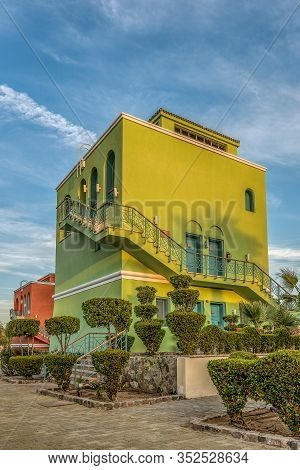 A Quadrangular Three-story Green House With An Outside Staircase In The Sunset Against A Blue Sky, E