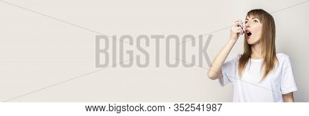 Young Woman With An Open Mouth In Her Hand Holds An Inhaler And Atomizes On A Light Background. Bann