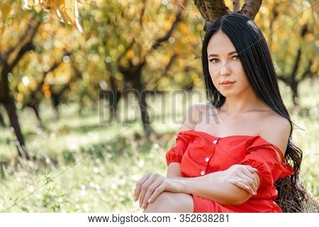 Walk In The Garden. A Brunette Woman In A Red Dress Sits On The Grass, Leaning On A Tree Trunk, And 
