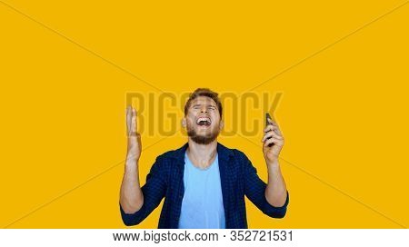 Bearded Caucasian Man Using A Phone Is Gesturing Up After Winning Something On A Yellow Wall