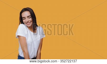 Delight Caucasian Girl Is Smiling With Closed Eyes Posing In A White T-shirt On A Yellow Wall With F