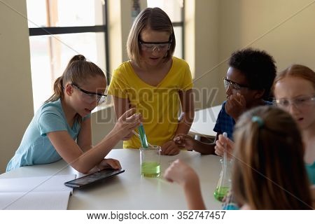 Front view of a multi-ethnic group of elementary school girls sitting at a table wearing safety glasses and using pipettes and beakers to do an experiment during a chemistry lesson, one girl holding a
