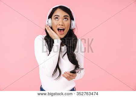 Cheerful pretty young asian woman listening to music with wireless headphones isolated over pink background, holding mobile phone