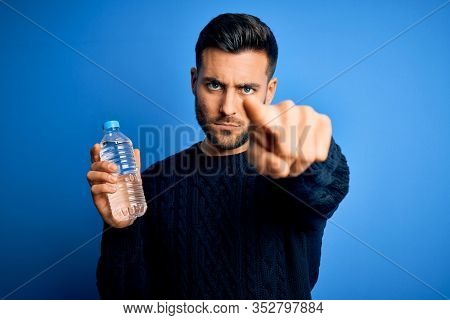 Young handsome man drinking bottle of water to refeshment over blue background pointing with finger to the camera and to you, hand sign, positive and confident gesture from the front